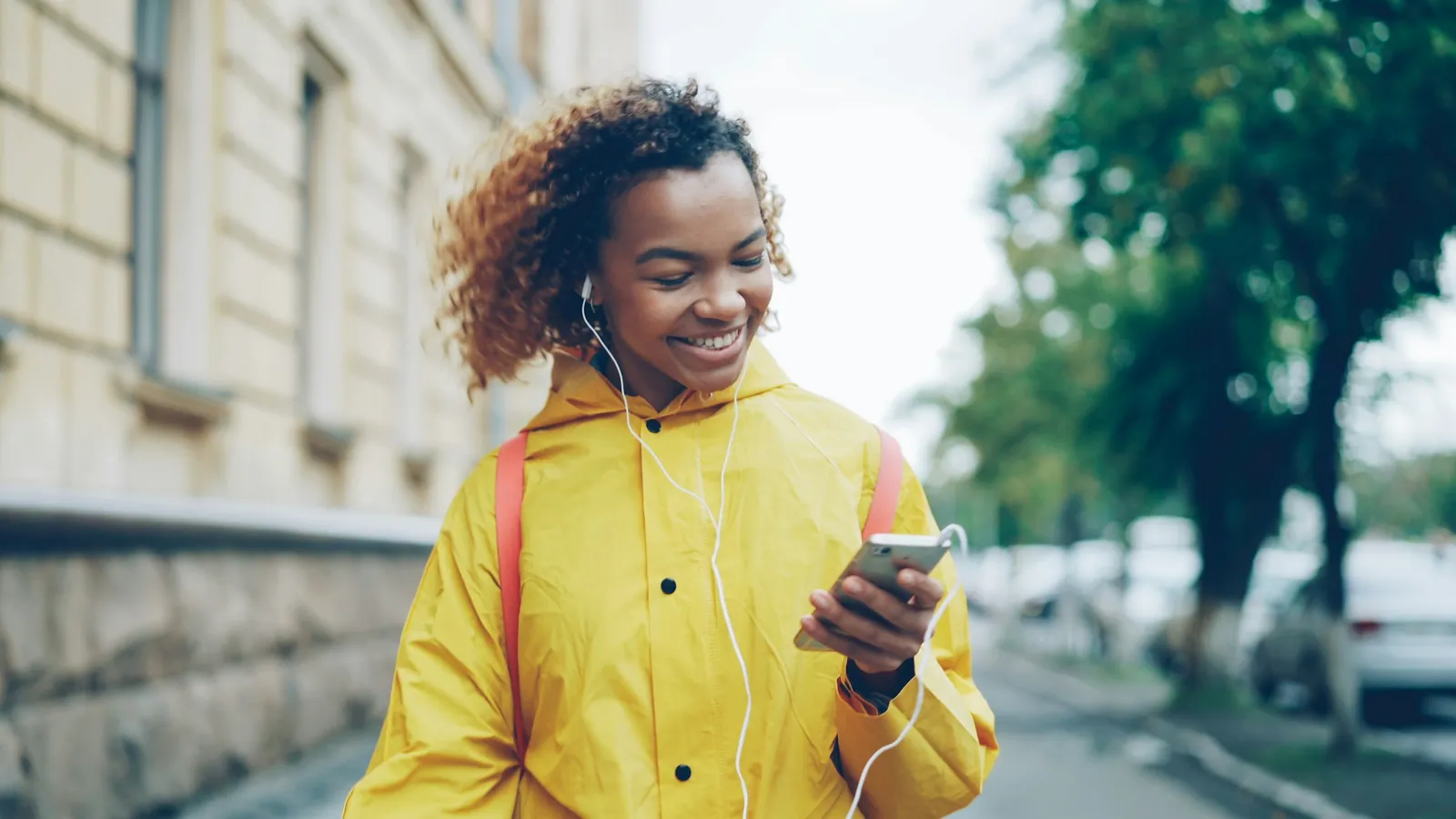 A young woman in a yellow raincoat walks down a city street with earphones in, smiling at her phone.